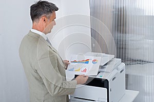 Businessman Looking At Paper With Graphs By Color Printer