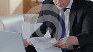 Businessman looking through papers and typing on laptop, statistical report