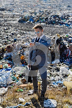 Businessman looking at watch, checking time standing on landfill, large pile of waste. Consumerism versus pollution