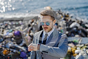 Businessman looking at watch, checking time standing on pile of waste on beach. Consumerism versus pollution concept