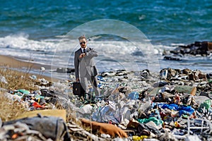 Businessman looking at watch, checking time standing on pile of waste on beach. Consumerism versus pollution concept