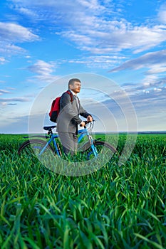 A businessman looks into the distance, he is standing with a backpack and a bicycle on a green grass field, dressed in a business