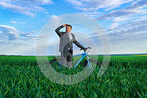 A businessman looks into the distance, he is standing with a backpack and a bicycle on a green grass field, dressed in a business
