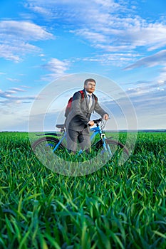 A businessman looks into the distance, he is standing with a backpack and a bicycle on a green grass field, dressed in a business