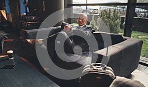 Businessman looks at his wristwatch waiting for meeting while sitting on large sofa in business center. Business travel