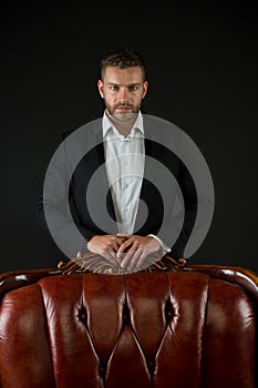 Businessman or man in formal suit on dark background. Man on serious face posing behind leather armchair. Business