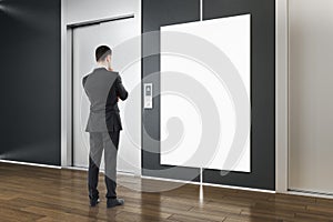 Businessman in modern concrete interior with elevators, empty white mock up poster and wooden parquet flooring. Lift, lobby and