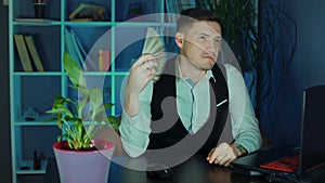Businessman with money at workplace. Young man waves money as fan, sitting in armchair before computer in office.