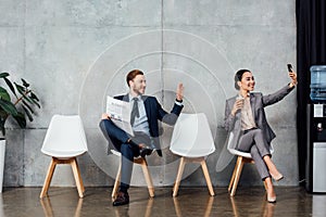 Businessman with newspaper waving with hand while asian businesswoman taking selfie on smartphone in