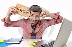 Businessman at office desk working on computer laptop asking for help holding cardboard sign looking sad and depressed