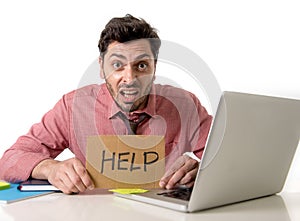 Businessman at office desk working on computer laptop asking for help holding cardboard sign looking sad and depressed