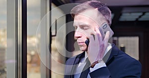 Businessman phone conversation. A young man stands in a gray suit in the office near the window and speaks on the phone