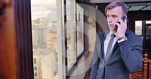 Businessman phone conversation. A young man stands in a gray suit in the office near the window and speaks on the phone