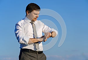 Businessman posing in a field, he looks at his wristwatch, green grass and blue sky as background