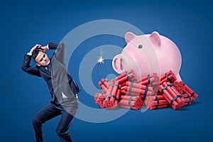 Businessman protecting himself with hands and pink piggy bank over red dynamite sticks with lit fuse on blue background