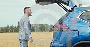 Businessman putting his sport bag into car trunk and closing it by pushing the button, countryside shot