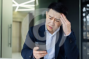 Businessman reading bad news online from phone, asian man disappointed and sad looking at smartphone screen, man working