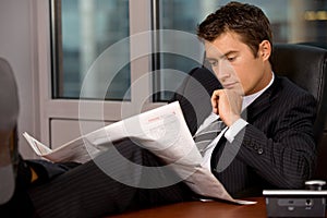 Businessman reading newspaper in office with hand on chin