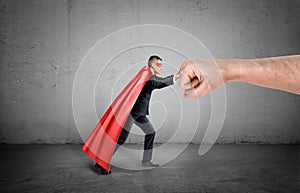 A businessman in a red cape and a mask pushing at a giant human fist on concrete background.