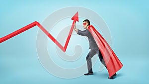 A businessman in a red flowing cape trying to bend a red statistic arrow upwards on blue background.