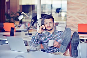 Businessman relaxing with legs up on desk, drinking coffee while dreaming about future at workplace in modern office.