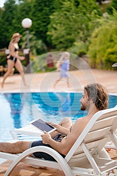 Businessman resting on sunbed near swimming pool while working remotely at laptop computer connected to wireless