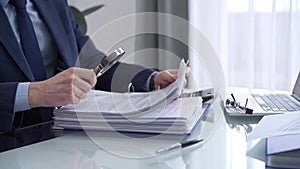 Businessman reviewing ring folder of financial documents with magnifying glass at desk in modern office. Audit and taxes