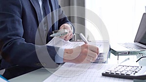 Businessman reviewing ring folder of financial documents with magnifying glass at desk in modern office. Audit and taxes