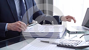 Businessman reviewing ring folder of financial documents with magnifying glass at desk in modern office. Audit and taxes