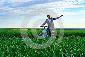 Businessman rides a bicycle through a green grass field and waves his hands, dressed in a business suit, beautiful nature in