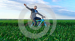 Businessman rides a bicycle through a green grass field and waves his hands, dressed in a business suit, beautiful nature in