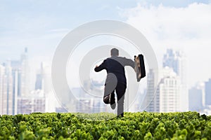 Businessman running towards the city with a briefcase in a green field with plants