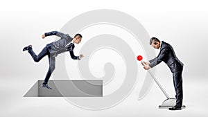 A businessman runs on white ground while a square hole controlled by a man on a lever opens right under his feet.