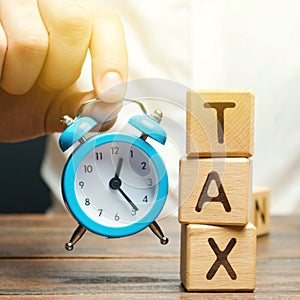 Businessman`s hand holding a clock near the wooden blocks with the word Tax. Time to pay taxes. The concept of annual taxation. photo