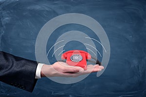 A businessman`s palm hold a small red retro dial phone with lines showing soundwave emissions.