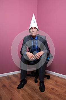 Businessman sat in corner wearing dunce hat