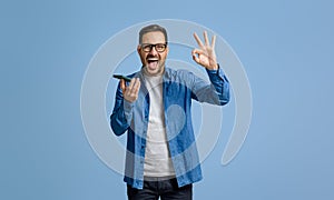 Businessman screaming ecstatically and showing OK sign while using cellphone on blue background