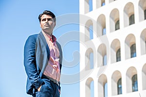 Businessman in shirt and jacket outdoors. Hands in the pocket. Behind him a building and the blue sky. Look into the distance