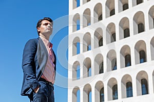 Businessman in shirt and jacket outdoors. Hands in the pocket. Behind him a building and the blue sky. Look into the distance