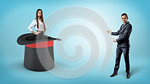 A businessman shows a self-assured businesswoman standing inside a giant illusionists hat on a blue background.