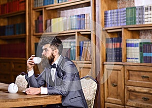 Businessman sits in vintage interior, holds book and cup of tea. Bearded man in luxury suit in his cabinet. Mature man with calm f