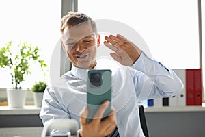 Businessman sits at workplace and waves his hand to smartphone in greeting.