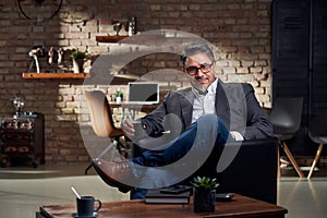 Businessman sitting in armchair in cosy home office, smiling, drinking coffee.