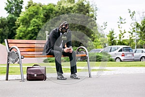 Businessman sitting on a banch wearing a gas mask with phone in hand