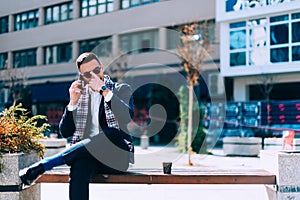 Businessman is sitting on bench in the city center and talking on his mobile