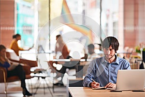 Businessman Sitting At Desk Writing In Notebook In Modern Open Plan Office