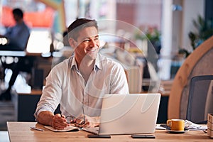 Businessman Sitting At Desk Writing In Notebook In Modern Open Plan Office