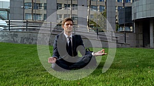 Businessman sitting in lotus position, meditating on lawn near office center