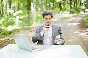 Businessman sitting at the office desk with laptop computer and cup of coffee use mobile phone in green forest park. Business conc