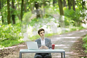 Businessman sitting at the office desk work at laptop computer and cup of coffee typing mobile phone on road of green forest park.